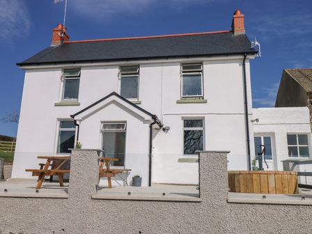 A white two-story house with a slate roof a wooden picnic table and a wooden circular structure on the patio at Llanwern Farm in Fishguard