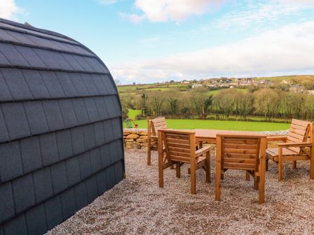 An outdoor seating area with wooden chairs and table next to a dome-shaped structure with a tiled exterior and a view of green fields and houses at Audrey in Penzance near Newlyn