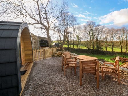 An outdoor seating area with wooden chairs and table on gravel near a wooden gate and curved wooden pod at Lloyd in Penzance near Newlyn