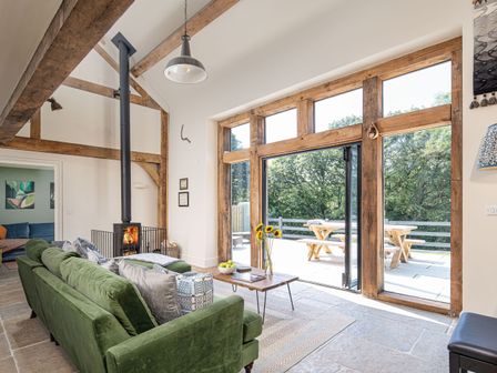 A living room with a green sofa coffee table wood burning stove and large wooden framed windows opening to a patio with outdoor seating at Ty Tylluan in Camrose