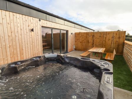 An outdoor area with a hot tub wooden picnic table and benches next to a building with sliding glass doors at Bears Court Barn 4 Little Rissington near Bourton-On-The-Water