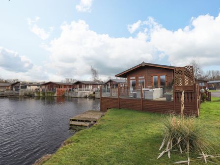 A wooden cabin with a deck and outdoor furniture beside a lake with other similar cabins nearby at Otters Reach near Carnforth
