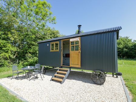 An outdoor area with a black corrugated metal shepherd hut on wheels and a small table with three metal chairs on gravel at Blackthorn in South Leigh near Witney
