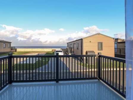 A view from a balcony overlooking a driveway with a car parked and a wooden cabin with the sea in the background at The Sea Lodge in Bacton near Walcott