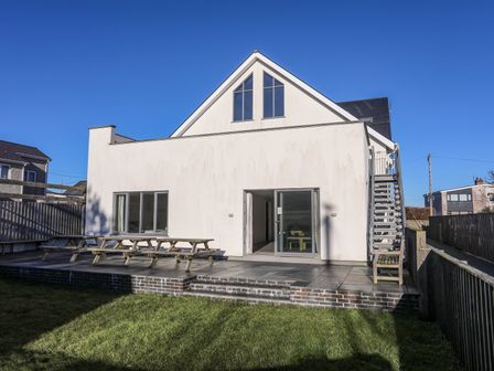The back of a white house with large windows and sliding doors opening to a patio with picnic tables and stairs to the upper level at Plas Gwylan in Rhosneigr
