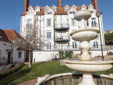 A large white multi-story building with balconies in the background and a stone water fountain in the foreground at Apartment 7 Mansion House in Tenby