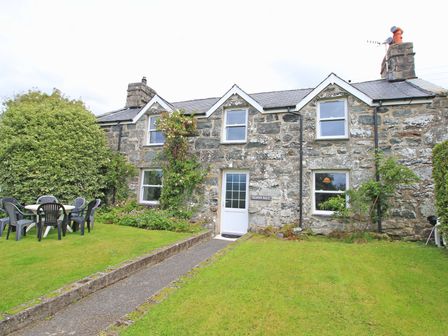 A stone cottage with a white door and four windows a garden with grass and outdoor chairs at The Farmhouse in Llanbedr