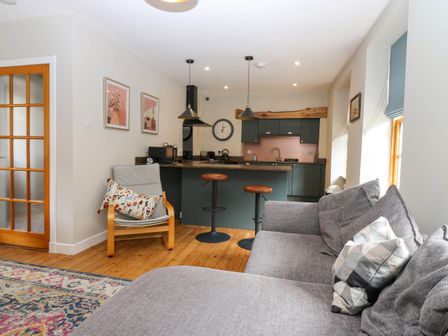 A living room with a grey sofa and armchair next to a kitchen with bar stools at 49 Castlegate in Jedburgh