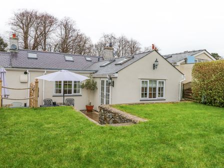 A garden with green grass and patio furniture with umbrellas outside a white cottage with multiple windows at Bramley Cottage in Beaumaris