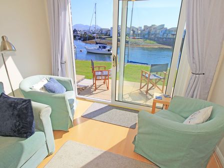 A sitting area with two armchairs and a sofa facing sliding glass doors overlooking a river with boats at Harbour Escape in Porthmadog