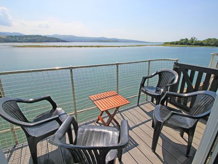 A balcony with four black plastic chairs and a small wooden table overlooking water and distant hills at Harbour Flat - 12E South Snowdon Wharf in Porthmadog