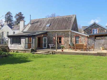 A stone house with a wooden patio and garden furniture on a lawn at The Old Coach House in Little Dunley Cottages near Bovey Tracey