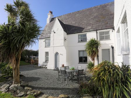 A white house with a slate roof and outdoor seating on a gravel patio with palm trees at School House in Aberffraw