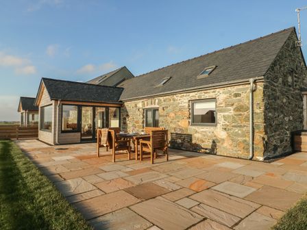 An outdoor patio with a wooden dining table and chairs next to a stone building at Cuddfan in Llanfwrog near Bodedern