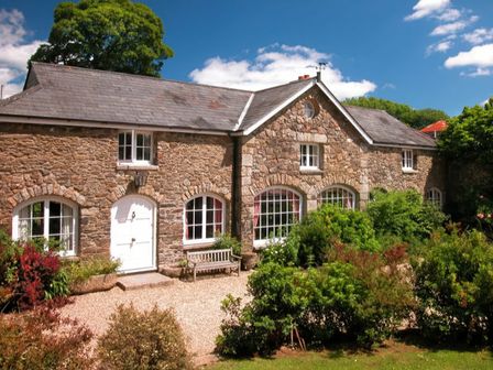 A stone house with white framed windows a white door a wooden bench and garden greenery at The Coach House in Lydford