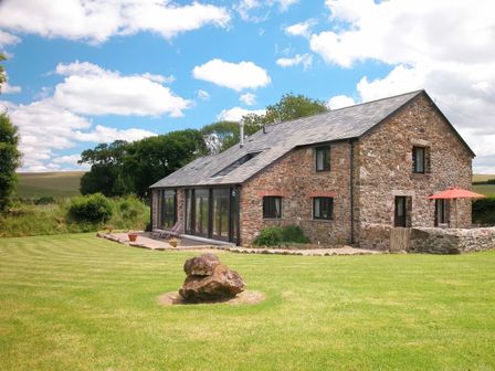 A stone cottage with glass doors and patio chairs on a lawn with a large rock and umbrella at The Red Barn in North Brentor near Lydford