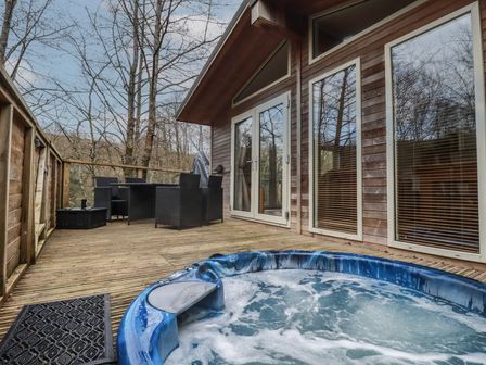 An outdoor deck with a hot tub black chairs and table next to a wooden building with large windows at 1 Miller's Island near Lanreath