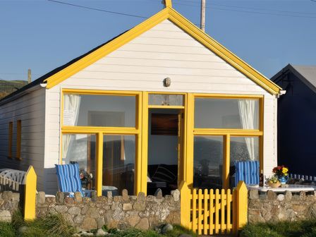 A small white house with yellow window frames and a yellow gated fence with blue striped chairs outside at Homestead in Aberdesach near Penygroes