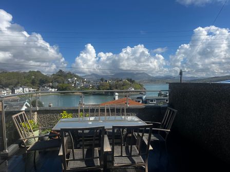 An outdoor patio with a glass table and chairs overlooking a body of water and houses at The Quarterdeck in Borth-Y-Gest