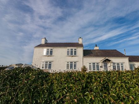 Exterior view of a white house with multiple windows and a hedge in the foreground at Fferm Porthdafarch South Farm House in Porthdafarch near Trearddur Bay