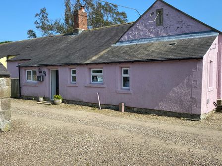 A pink single-story house with multiple windows and a chimney on a gravel driveway at The Smithy in Chathill near Seahouses
