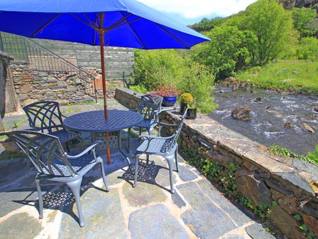 An outdoor patio with metal chairs and a table under a blue umbrella next to a river at Riverbank Cottage in Beddgelert