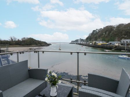 A balcony with woven chairs and a table with flowers overlooking a river with boats and trees at Anchor Watch in Porthmadog