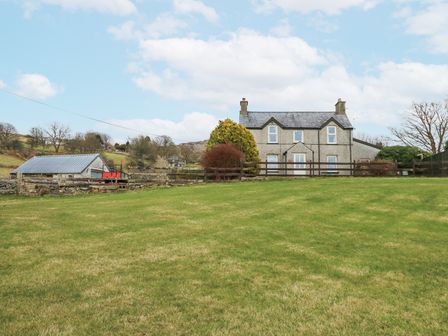 A stone house with two chimneys surrounded by grass and a wooden fence at Dalar Cottage in Garndolbenmaen near Criccieth