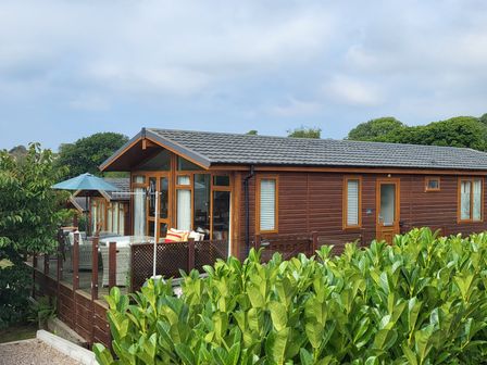 A wooden cabin with a fenced patio and outdoor furniture surrounded by green plants at Matilda Lodge in Newton Ferrers