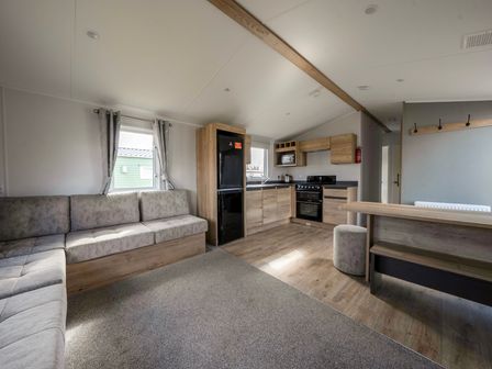 An open living space with a sofa, kitchen area, black refrigerator, oven, and dining bench at Kingfisher Caravan - Billing Aquadrome Holiday Park