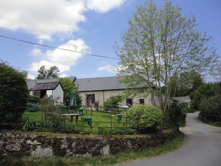 A stone building with a slate roof beside a road and a garden with green chairs and a table at Moorhaven in Chagford