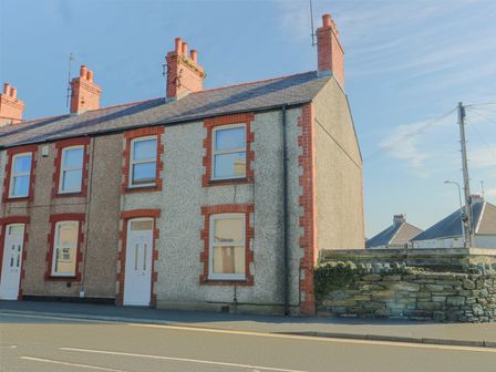 A two-story end terrace house with red brick chimneys and window frames and a white door at 63 Kingsland Road in Holyhead