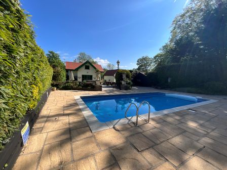 An outdoor swimming pool with patio and a two story house with red roof in the background at Hafod Lodge in Hensol near Pontyclun