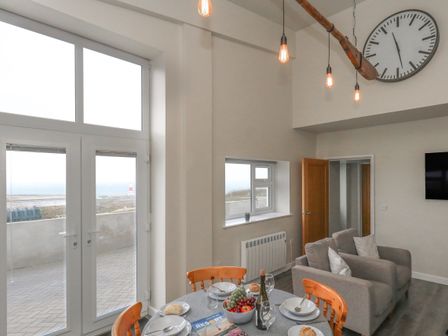A dining and living area with a round table set for four near large windows and a glass door overlooking an outdoor area with a lighthouse visible beyond at Coastguard View in Portland Bill