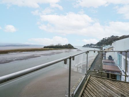 A balcony with wooden flooring and glass railing overlooking a river and an island at Harbour Apartment in Porthmadog