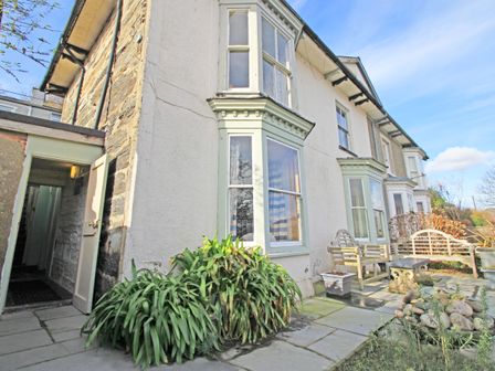 An exterior view of a house with bay windows a patio with wooden benches a table potted plants and greenery at Garth Cottage in Porthmadog