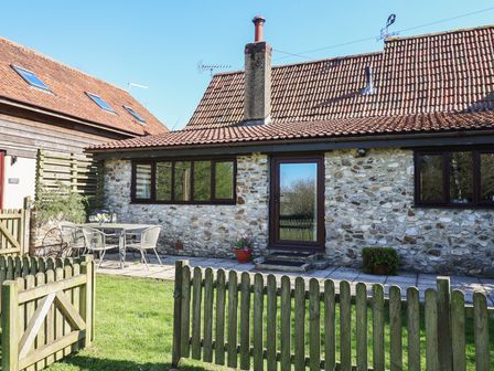 A stone cottage with a tiled roof a small patio with a table and chairs and a wooden picket fence at Weathervane Cottage in Upottery