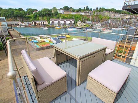 An outdoor seating area with cushioned wicker furniture on a balcony overlooking a marina with boats and buildings in the background at Dros Y Mor in Porthmadog