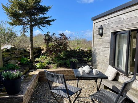 A small outdoor seating area with a table and two chairs beside a wooden building and garden at The Cow Shed in Porthcothan near Mawgan Porth