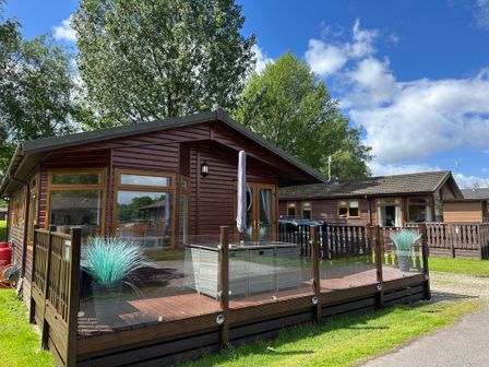 A wooden cabin with glass railing and outdoor seating on a deck surrounded by trees at Lakeside 18 in Carnforth
