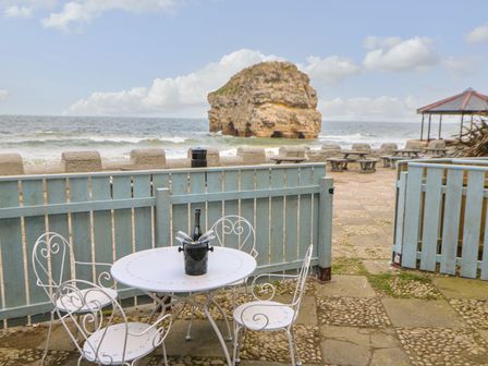 An outdoor patio with a white metal table and chairs and a bottle on the table overlooking the sea and a large rock at The Beach Apartment in South Shields