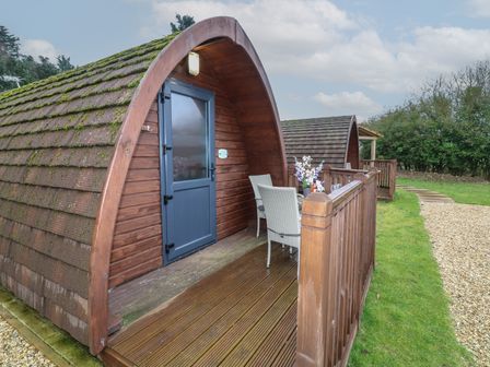 Two wooden pods with blue entrance doors and outdoor seating on a wooden deck near a gravel path and grassy area at Chantelles in Holbeach