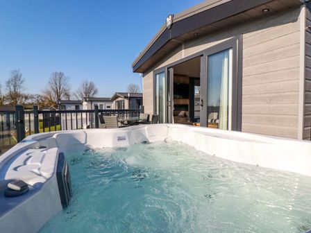 An outdoor hot tub on a deck next to a building with sliding glass doors and outdoor furniture at The Maples No 48 in Hewas Water near Sticker
