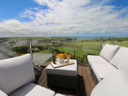 An outdoor seating area with white cushioned chairs and a table with drinks and flowers overlooking fields and the sea at Coastmans Nest in Mawgan Porth