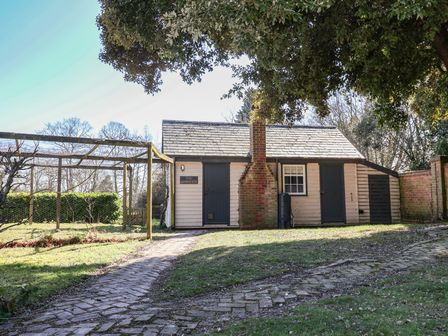 A small wooden building with a chimney and two doors beside a brick path and a fenced garden area at The Woodshed in Tendring near Weeley