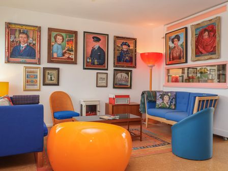 A living room with blue furniture orange chair paintings on walls and a floor lamp at The Orchard Close Art House Great Livermere near Bury St Edmunds