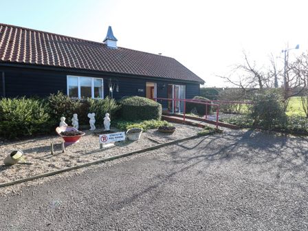 A building with a tiled roof and dark exterior with a ramp leading to the entrance surrounded by bushes and garden ornaments at Swallow Barn in Holton near Halesworth