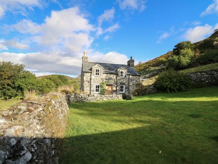 A stone cottage with a slate roof surrounded by a stone wall and green grass at Tyn Y Graig in Fairbourne