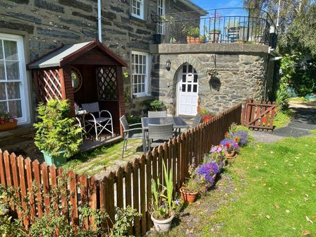 An outdoor patio area with a small wooden gazebo chairs a table and potted flowers behind a wooden fence at Garden Flat in Porthmadog
