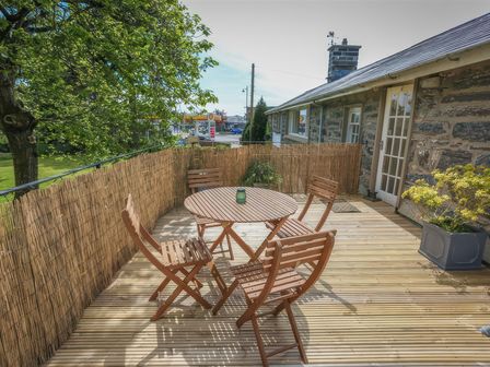 A wooden outdoor patio with a round table and four chairs near a stone building at Treetops Flat in Porthmadog